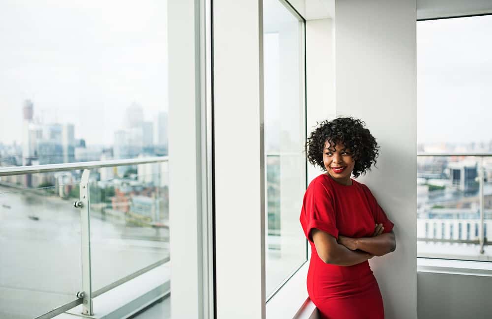  A woman in a red dress standing in front of a window, looking out at the city. She has her arms crossed and is smiling.